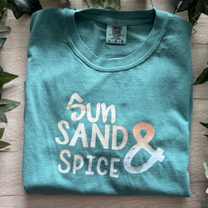 A folded teal t-shirt with a distressed graphic reading 'Sun SAND & SPICE' in white and orange, styled on a light wood background with green ivy leaves.