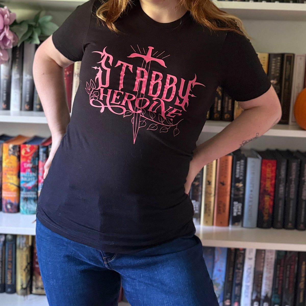A woman with light brown hair stands in front of white bookshelves filled with books, wearing a black graphic t-shirt with pink text that reads 'STABBY Heroine' and blue jeans.